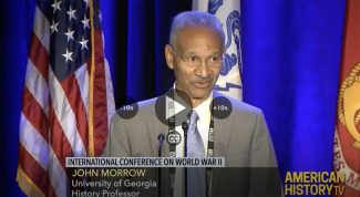 photo of man speaking at microphone, with flags and lower thirds text