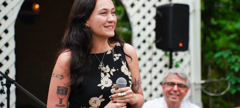 photo of woman with microphone, man in background, outdoors