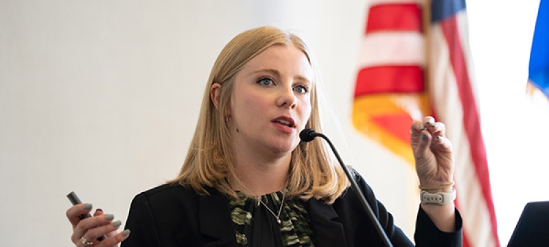 photo of woman speaking at lectern, flags in background