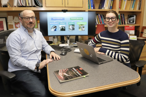 photo of two people sitting across a table, computer screen and books in background